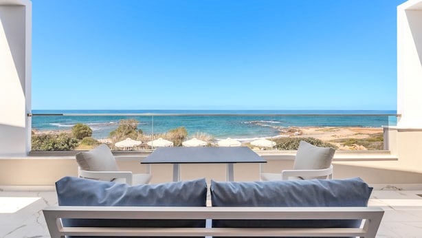 A balcony with a table and two chairs, overlooking a beach and ocean. - Elmar bay Villa (Hersonissos)