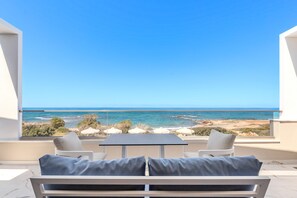 A balcony with a table and two chairs, overlooking a beach and ocean. - Elmar bay Villa (Hersonissos)