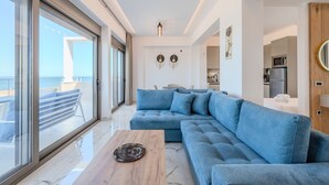A modern living room with a large blue sectional sofa, a wooden coffee table, and a view of the sea through large windows. - Elmar bay Villa (Hersonissos)