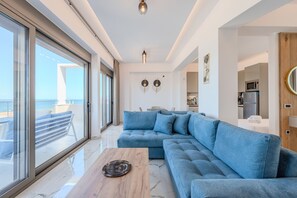 A modern living room with a large blue sectional sofa, a wooden coffee table, and a view of the sea through large windows. - Elmar bay Villa (Hersonissos)
