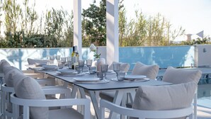 A dining table set with plates, glasses, and bottles, surrounded by white chairs with cushions, overlooking a pool. - Elmar bay Villa (Hersonissos)