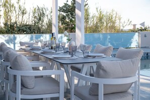 A dining table set with plates, glasses, and bottles, surrounded by white chairs with cushions, overlooking a pool. - Elmar bay Villa (Hersonissos)