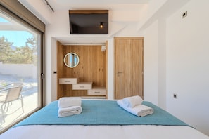 A modern bedroom with a bed, a TV, a wooden wardrobe, and a bathroom with a mirror and sink. - Elmar Bay Beachfront luxury Villa in Hersonissos (Limenas Chersonisoy)