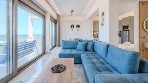 Una sala moderna con un amplio sofá seccional azul, una mesa de centro de madera y vista al mar a través de grandes ventanales.