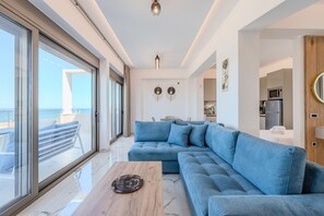 A modern living room with a large blue sectional sofa, a wooden coffee table, and a view of the sea through large windows. - Elmar Bay Beachfront luxury Villa in Hersonissos (Limenas Chersonisoy)