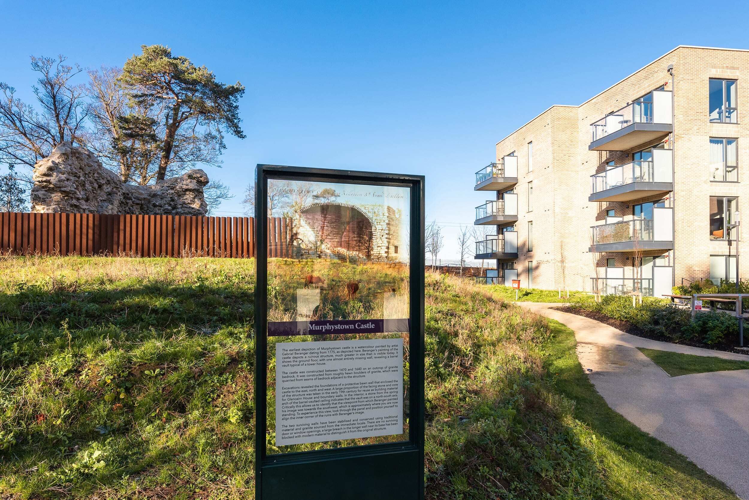 Un letrero con información histórica sobre un castillo, un área verde y un edificio moderno con balcones.