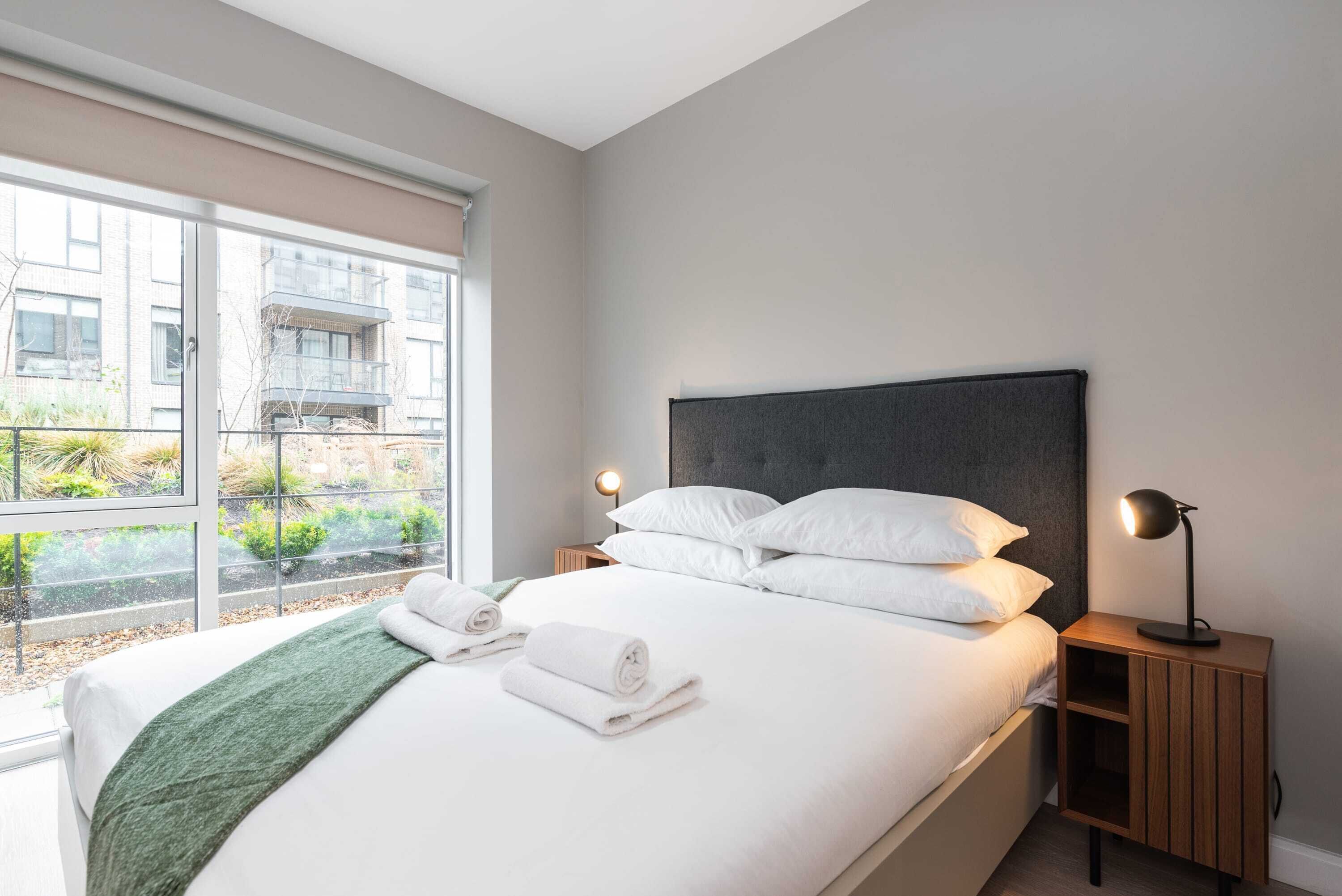 Una habitación de hotel moderna con una cama grande, dos mesitas de noche con lámparas, vistas amplias a través de un ventanal y una manta verde.