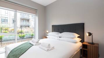 Una habitación de hotel moderna con una cama grande, dos mesitas de noche con lámparas, vistas amplias a través de un ventanal y una manta verde.
