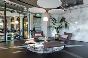 A modern lobby with a marble wall, round tables, leather sofas, and a large potted plant. - AEA-Woodward Square (Dublin)