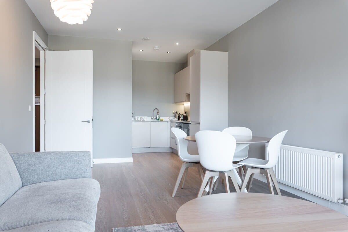 Un salon moderne avec un canapé gris, une table ronde en bois et des chaises blanches. On aperçoit à l’arrière-plan une cuisine avec des meubles blancs et un coin repas.