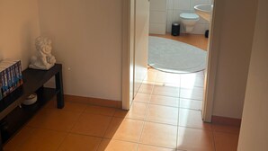 Een hal met een betegelde vloer, een witte deur die naar een badkamer leidt, en een klein tafeltje met een beeld en boeken.