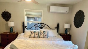 Um quarto com cama, duas mesas de cabeceira, janela com persianas, ar condicionado e arte decorativa na parede.