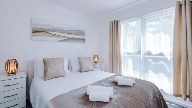 A hotel room with a large bed, two bedside lamps, a wall-mounted abstract painting, and a view through a window with curtains. - Central Nottingham 3 Bed House with Driveway (Nottingham)