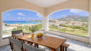 A room with a wooden table, chairs, a vase with fruit, and a view of the sea and town. - Elysian Seaside Villa (Almyrida)