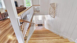 A wooden staircase with white railings, a view of a living room with a brown sofa, and a window with white curtains. - The Low Country Loft- Comfy 1BDRM Loft-4 Pass Golf Cart Included (Daufuskie Island)