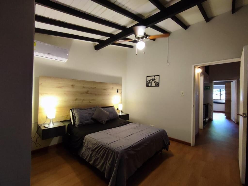 A bedroom with a bed, bedside lamps, a wooden headboard, a ceiling fan, and a hallway leading to another room.