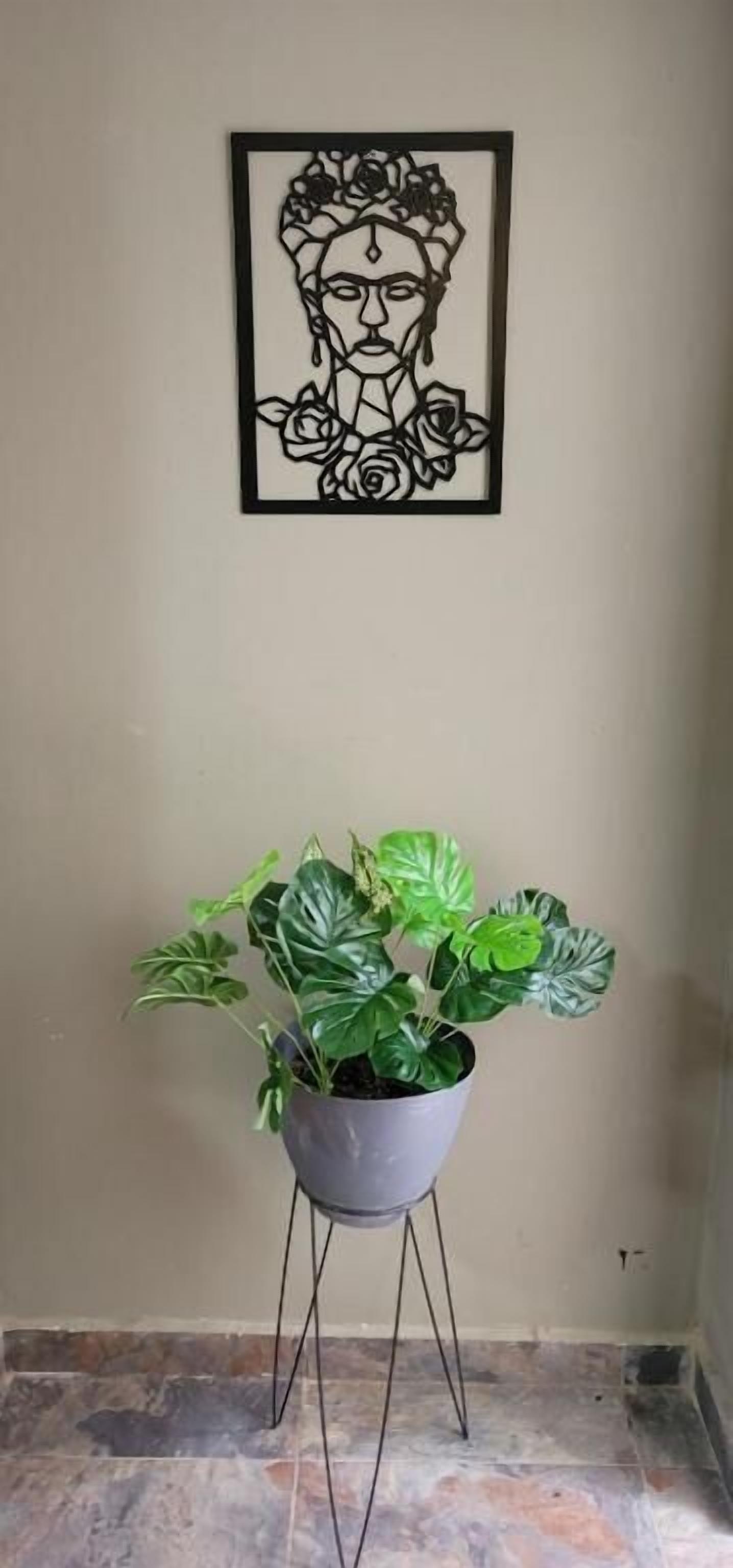 A framed artwork with a floral design on a wall, a potted plant with broad leaves on a stand, and a tiled floor.