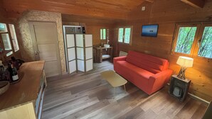 Ein Holzblockhaus-Interieur mit einer Couch, einem Tisch, einer Lampe und Blick auf den Wald durch Fenster.