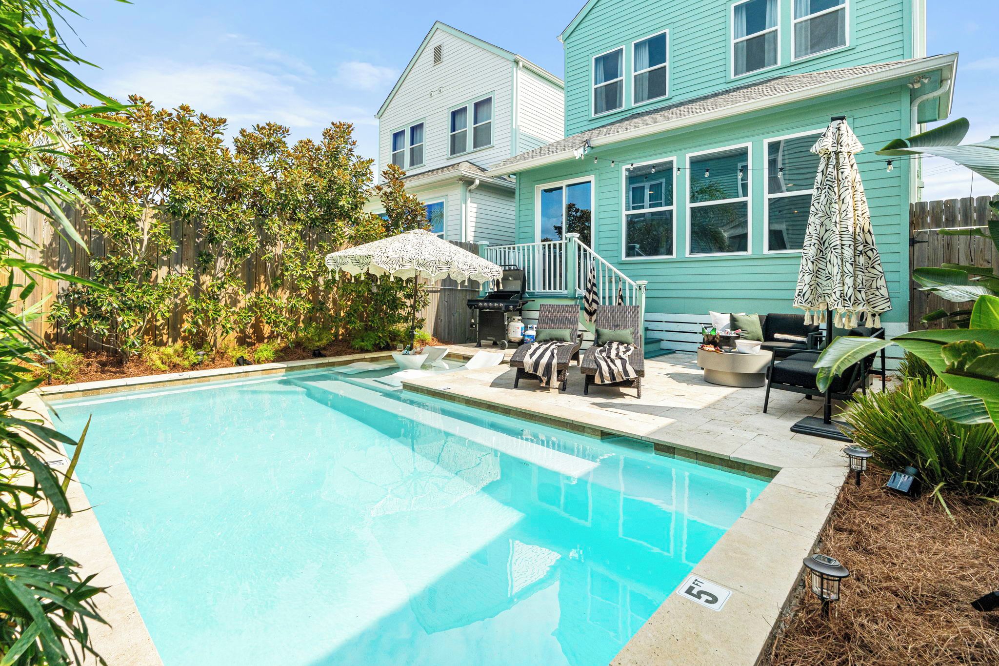 Un jardin avec une piscine, du mobilier d’extérieur et une maison dont le revêtement extérieur est de couleur turquoise.