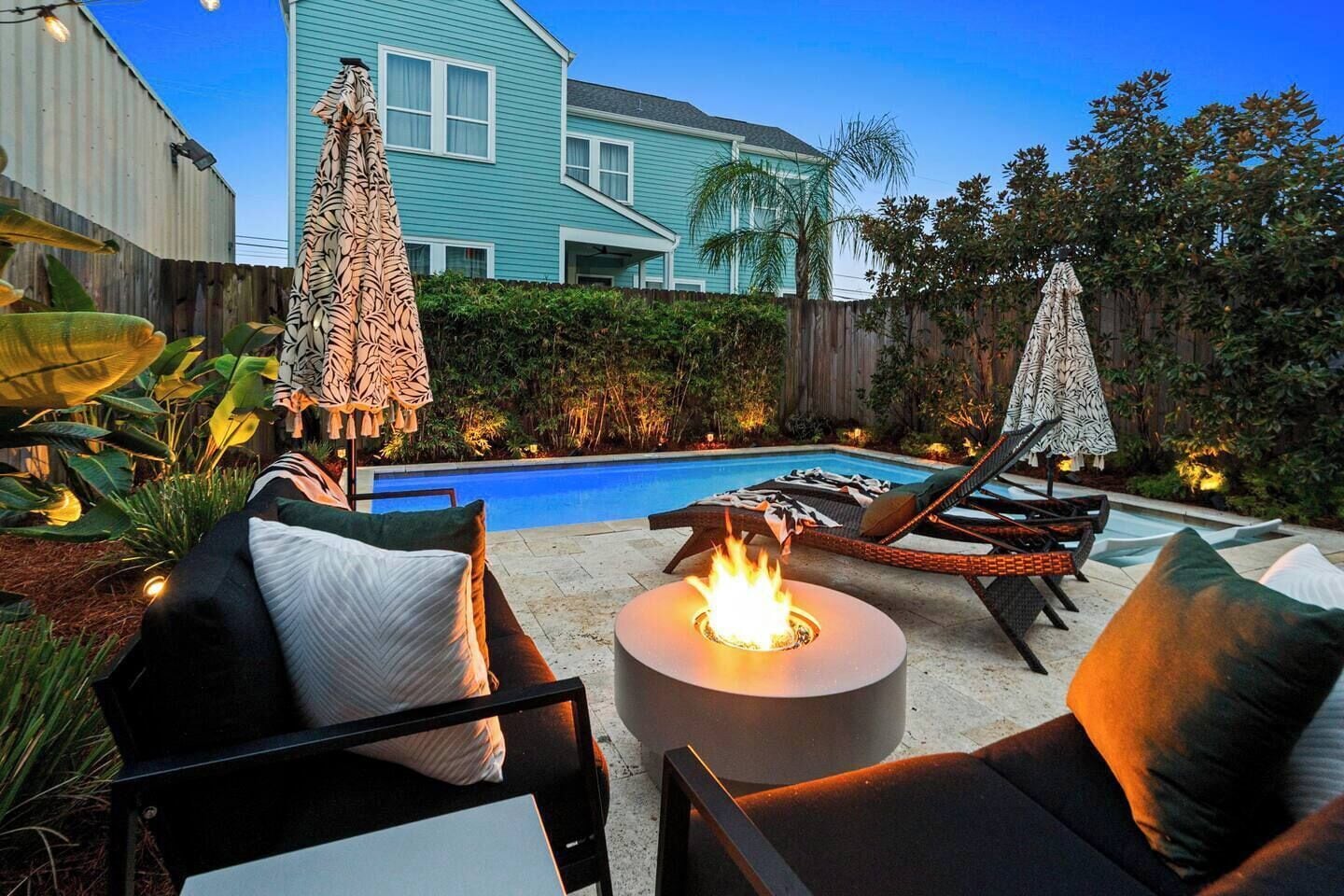 A backyard with a swimming pool, lounge chairs, a fire pit, and a house in the background.