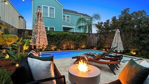 A backyard with a swimming pool, lounge chairs, a fire pit, and a house in the background.