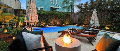 A backyard with a swimming pool, lounge chairs, a fire pit, and a house in the background.