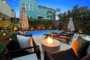 A backyard with a swimming pool, lounge chairs, a fire pit, and a house in the background.