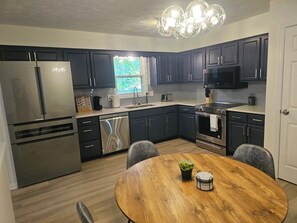 A modern kitchen with dark cabinets, stainless steel appliances, a wooden dining table, and a chandelier. - 3 Bedroom Grand Park Retreat in Westfield, IN (Westfield)