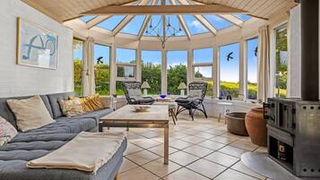 A conservatory with a wooden ceiling, large windows, a sofa, chairs, a table, and a wood stove.