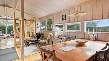 A spacious room with a wooden dining table, chairs, a bookshelf, and large windows.