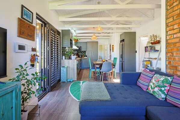A cozy living room with a blue sofa, a dining area with chairs, a kitchen with a counter, and a brick wall.