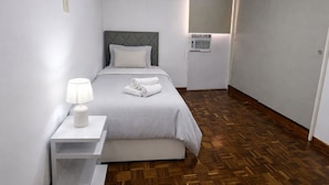 Un lit simple avec une tête de lit grise, une table de chevet avec une lampe et une serviette enroulée posée sur le lit.