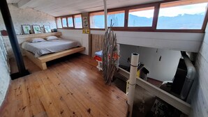 A bedroom with a bed, wooden floor, windows, and a view of mountains.