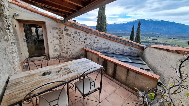 A rustic outdoor patio with a table and chairs, a stone wall, and a view of mountains.