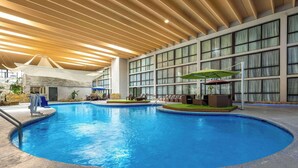 An indoor swimming pool with a wooden ceiling, large windows, and lounge areas with umbrellas.