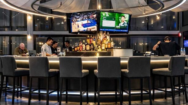 A modern bar area with multiple screens, a well-stocked bar, and a circular ceiling light fixture.