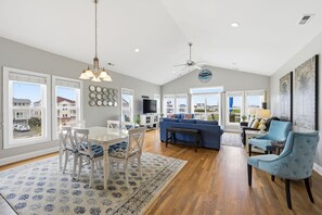 A spacious living room with a dining area, a blue sofa, and a ceiling fan. - Salt Life | Deck Views + Steps to Beach, Sleeps 10 (North Topsail Beach)