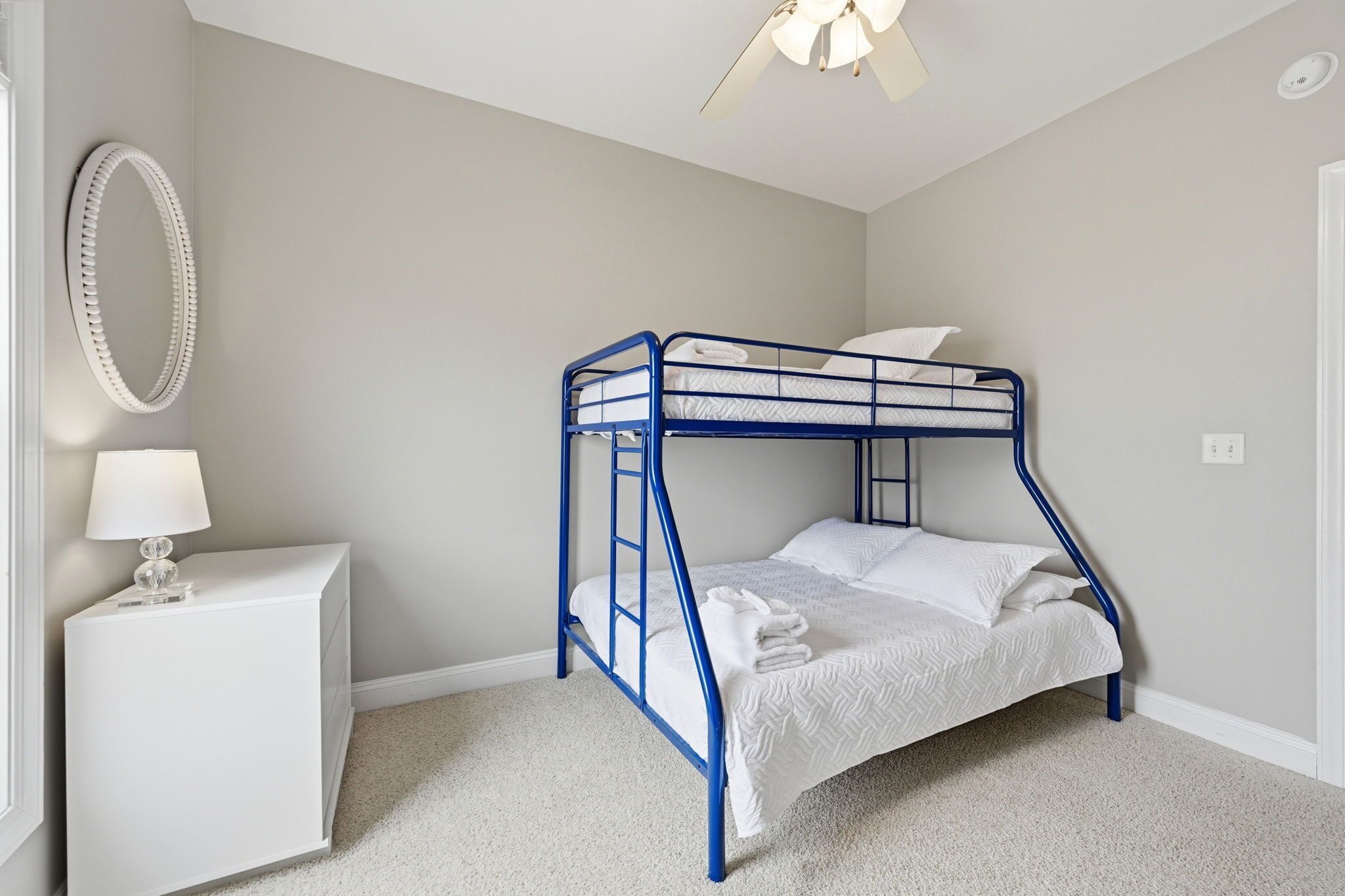 A bunk bed with a white mattress and pillows, a white bedside table with a lamp, and a wall-mounted mirror.