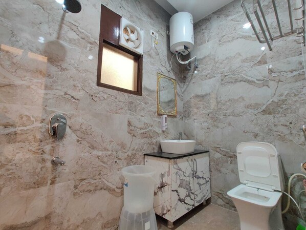 Casa de banho com parede de mármore, lavatório branco, sanita, espelho e ventoinha.