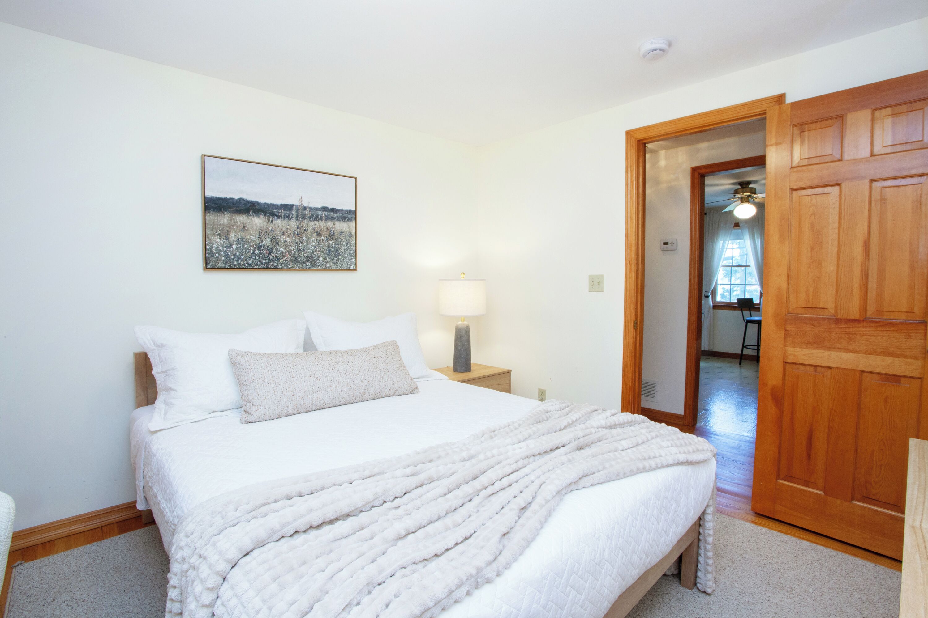 A bedroom with a bed, a nightstand with a lamp, a picture on the wall, and a wooden door.