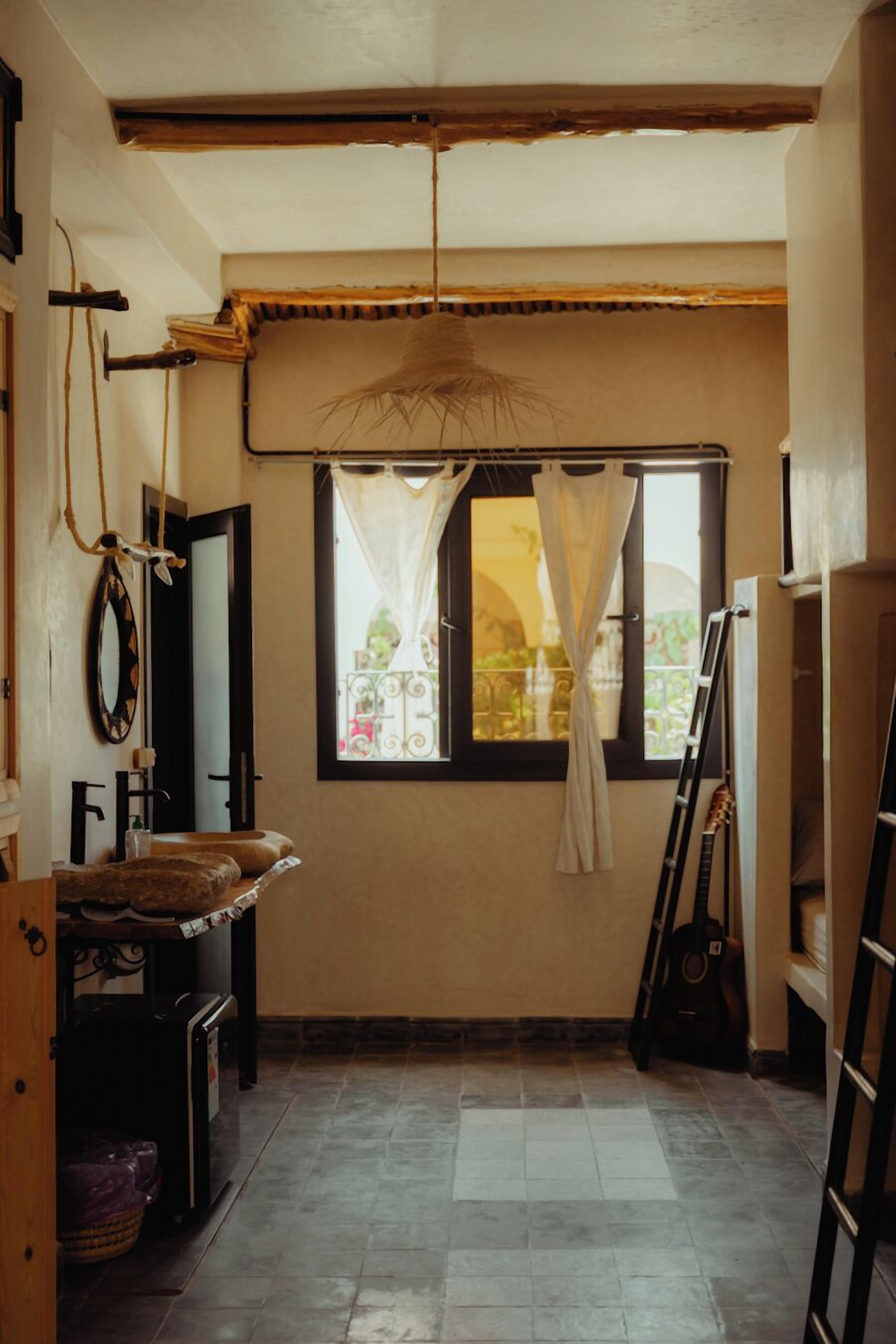 Una habitación con ventana, una guitarra apoyada en la pared, una escalera y una pequeña mesa con cojines.