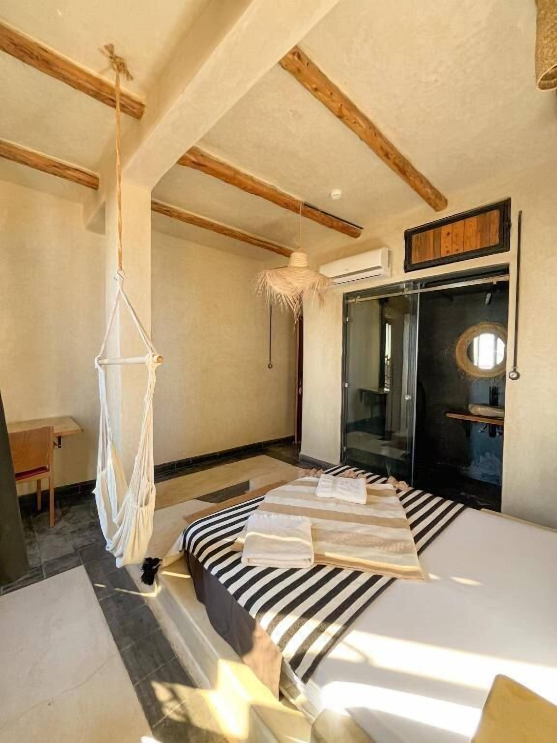 Un dormitorio con una cama, una silla colgante, un escritorio y una puerta de vidrio que da a un baño.