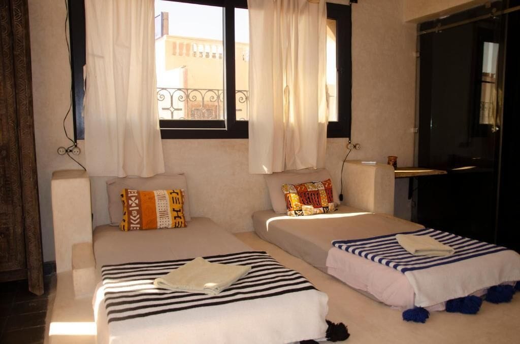 Una habitación con dos camas, una ventana con cortinas, una puerta y vistas a edificios.
