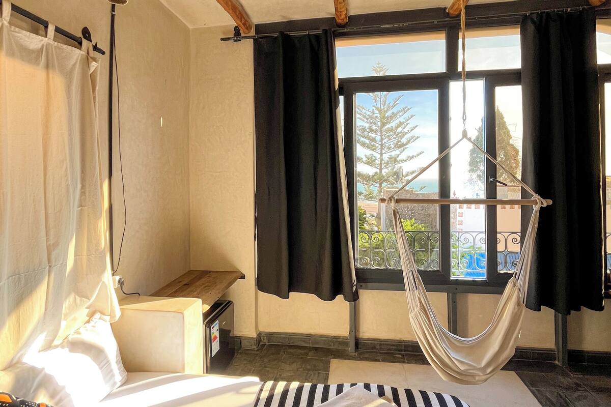 Een slaapkamer met een bed, een hangmat, een raam met gordijnen en uitzicht op de omgeving.