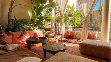 Uma sala de estar aconchegante com sofá, puf e mesa de centro, rodeada por plantas e cortinas.