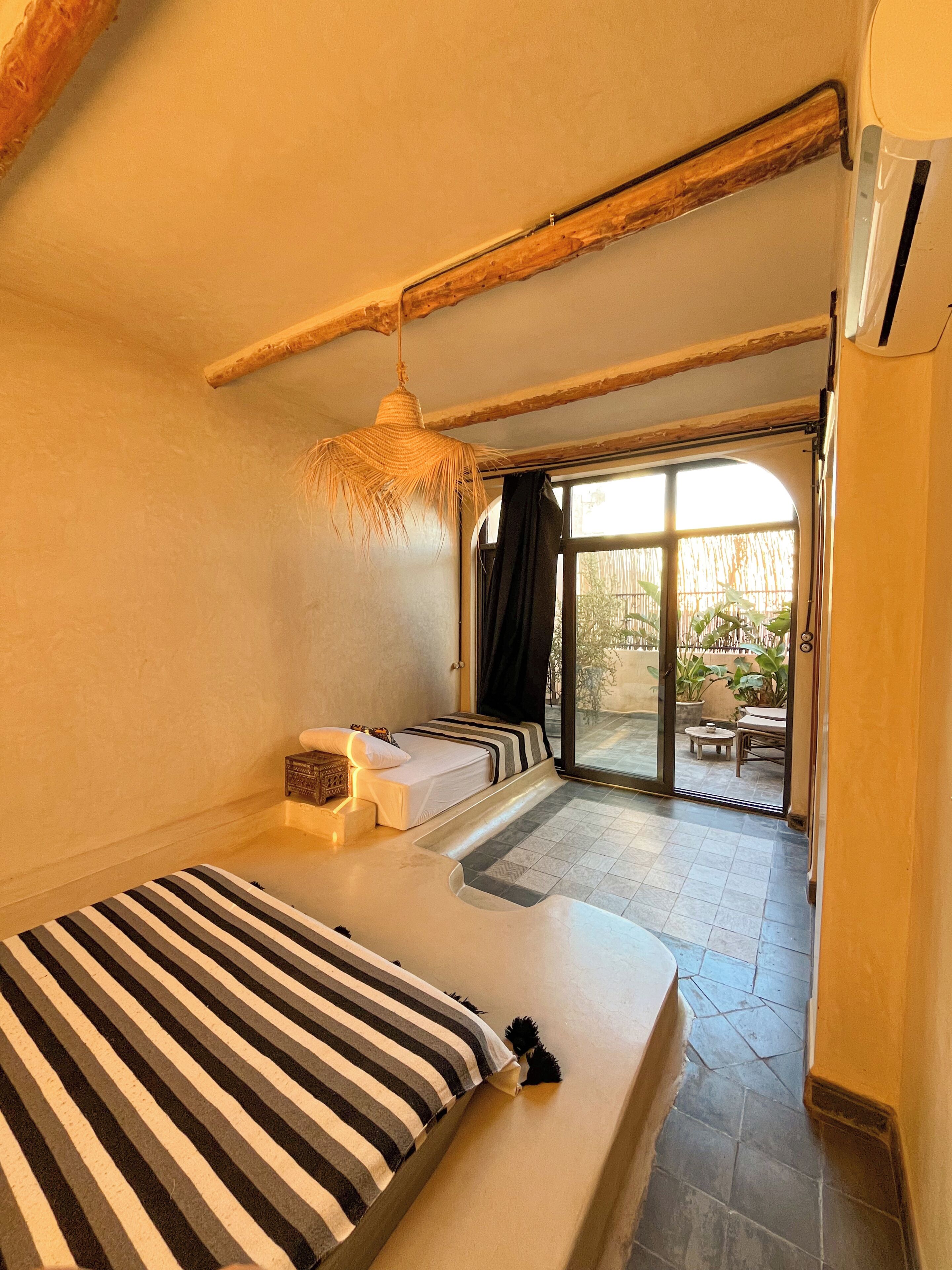 Een kamer met een bed, een klein tafeltje, een hanglamp en een uitzicht door een glazen deur op een buitenruimte.