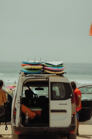 Una camioneta blanca con una baca transportando varias tablas de surf, estacionada en una playa con el océano al fondo.