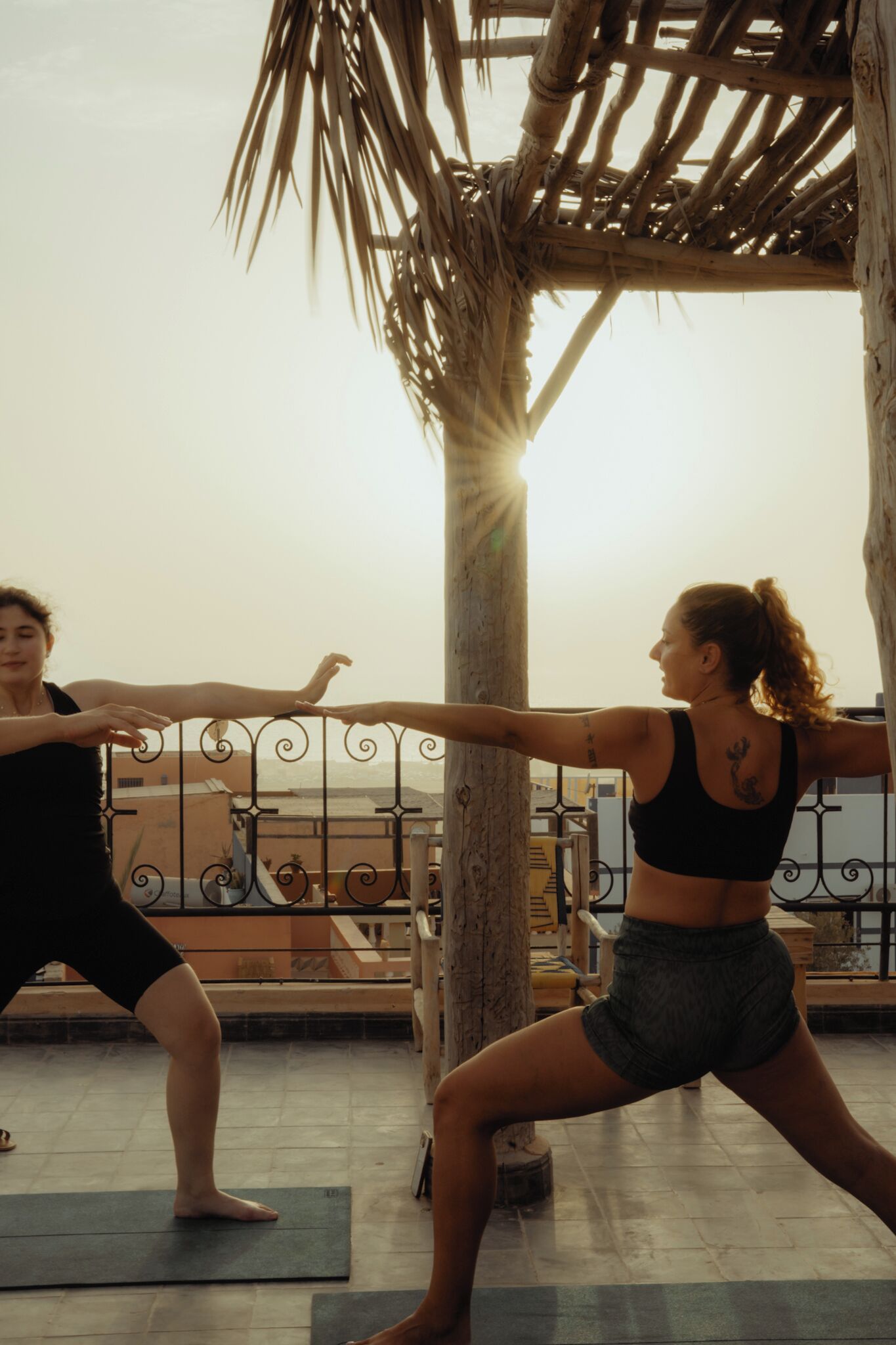 Two individuals practicing yoga on outdoor mats with a railing and palm tree in the background.