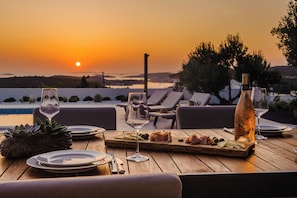 Ein Holztisch, gedeckt für eine Mahlzeit, mit Tellern, Besteck und Gläsern, mit Blick auf einen Sonnenuntergang und ein Gewässer.