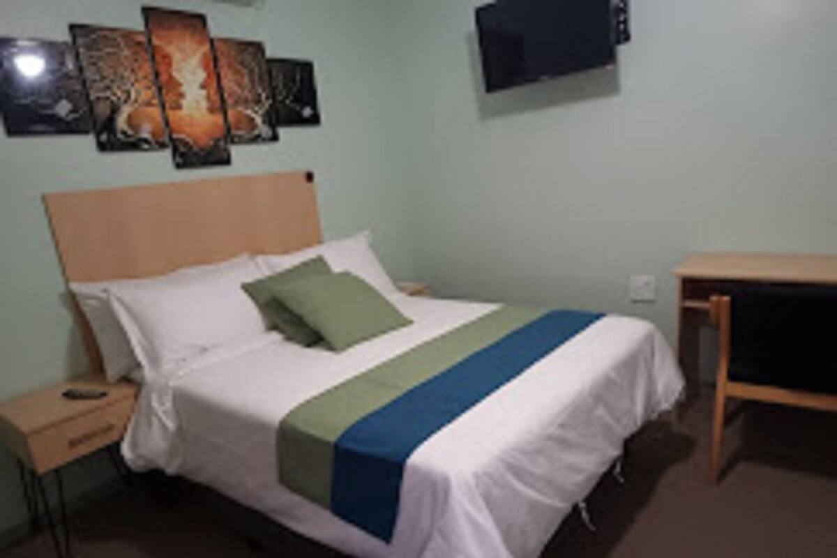 Un letto singolo con testiera in legno, un comodino, un cuscino verde e una televisione montata a parete.
